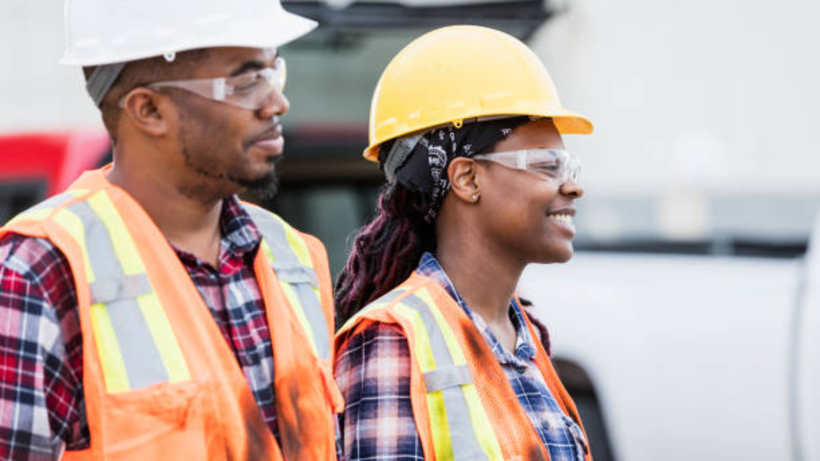 Two construction workers standing outdoors, wearing hard hats, safety glasses, reflective vest and plaid shirts. The focus is on the young African-American woman. Her coworker is in his 30s.