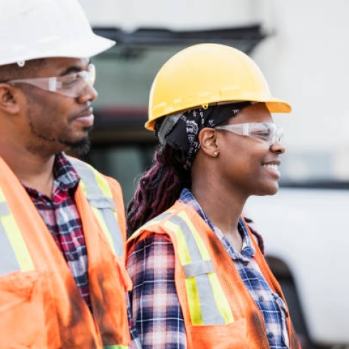 Two construction workers standing outdoors, wearing hard hats, safety glasses, reflective vest and plaid shirts. The focus is on the young African-American woman. Her coworker is in his 30s.